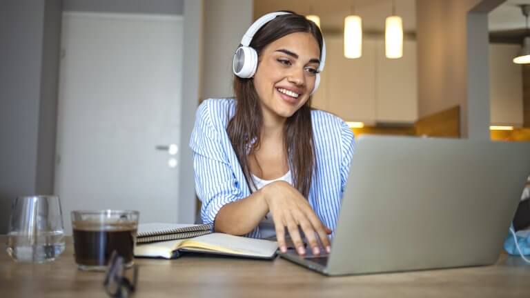 Smiling girl student wear wireless headphone study online with skype teacher, happy young woman learn language listen lecture watch webinar write notes look at laptop sit in cafe, distant education